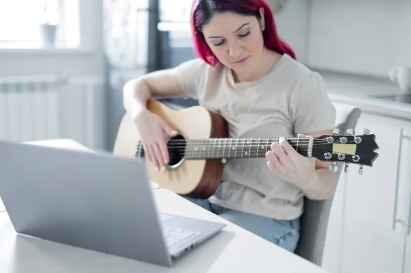 Eine Frau sitzt während einer Akustikgitarrenstunde in der Küche. Ein Mädchen lernt Gitarre spielen und schaut sich Lehrvideos auf einem Laptop an — Stockfoto