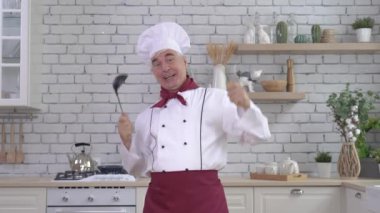 Dost canlısı, yaşlı bir şef aletlerini mutfakta kondüktör gibi sallıyor. Adam bir kepçe tutuyor ve çırpıyor.