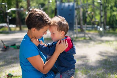 The woman gently hugs her son on the playground. The little boy is naughty and his mother calms him down clipart