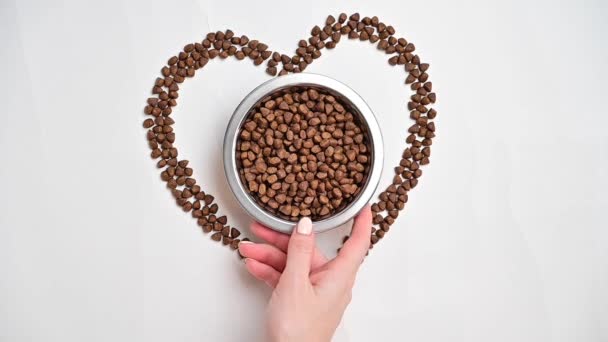 Dry food for dogs or cats is laid out in the shape of a heart on a white background. A woman puts a plate of food for pets — Stock Video
