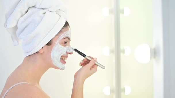 一个头上披着白毛巾的女人用刷子把蓝色黏土的面罩贴在脸上。照顾家里的皮肤 — 图库视频影像