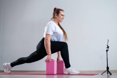 Evde spor egzersizleri yapan genç bir kadın. Kız evde paspasın üzerinde spor yapıyor ve cep telefonundan eğitim videosu izliyor.