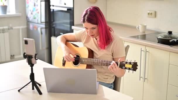Una mujer se sienta en la cocina durante una lección de guitarra acústica remota. Una chica aprende a tocar la guitarra y ve videos educativos en una computadora portátil — Vídeo de stock