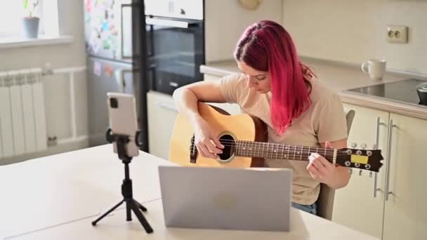 Een vrouw zit in de keuken tijdens een akoestische gitaarles op afstand. Een meisje leert gitaar spelen en kijkt educatieve video 's op een laptop — Stockvideo