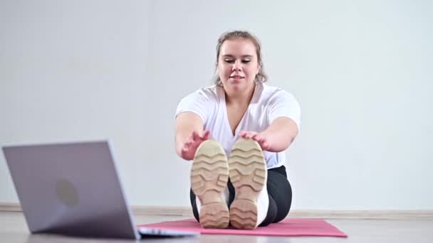 Joven mujer gorda caucásica haciendo curvas en una alfombra deportiva y viendo un video de entrenamiento en un ordenador portátil. Una chica gordita haciendo estiramiento de forma remota utilizando la comunicación de vídeo — Vídeo de stock