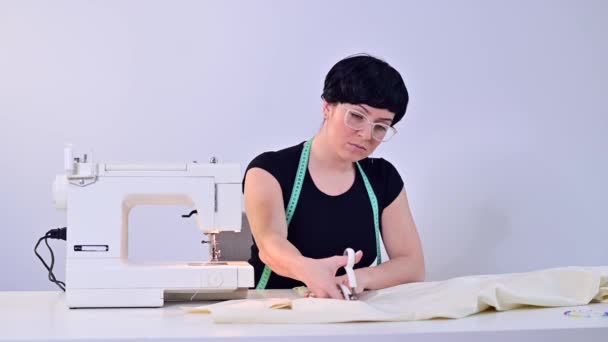 Професійна швачка вирізає матеріал ножицями на білому тлі. Жінка працює на швейній машині . — стокове відео