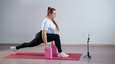 Genç, şişman, beyaz bir kadın spor paspasına basıyor. Çekici ve spor giyimli bir kız telefonda beyaz bir arka planda spor eğitmeni ile spor egzersizleri yapıyor.