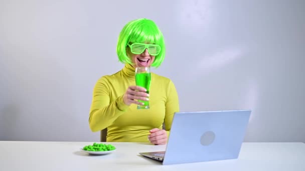 Wanita kaukasia dengan wig dan kacamata lucu minum bir hijau dan merayakan hari para patrick di rumah. Gadis berbicara dengan teman-teman melalui video komunikasi pada laptop. — Stok Video