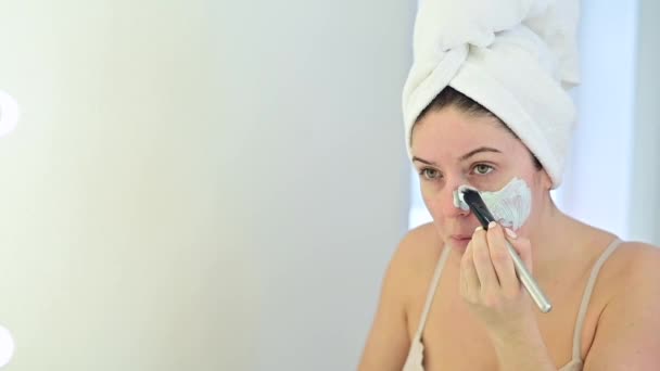Reflexão no espelho uma mulher em uma toalha branca em sua cabeça aplica uma máscara de barro azul em seu rosto com uma escova. Cuidar da pele em casa — Vídeo de Stock