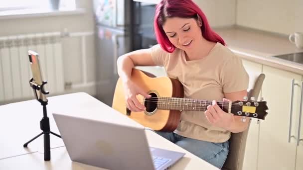 Una mujer se sienta en la cocina durante una lección de guitarra acústica remota. Una chica aprende a tocar la guitarra y ve videos educativos en una computadora portátil — Vídeo de stock