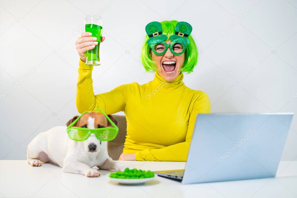 A young woman in a green wig and cheerful glasses drinks beer and bites glazed nuts. A girl sits with a dog at a table and celebrates st patricks day online chatting with friends on a laptop.