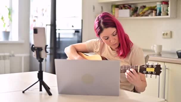 Seorang wanita duduk di dapur selama pelajaran gitar akustik jarak jauh. Seorang gadis belajar bermain gitar dan menonton video pendidikan di laptop — Stok Video