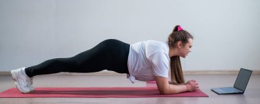 Genç, şişman, beyaz bir kadın spor paspası üzerinde tahta yapıyor. Spor kıyafetleri içinde yakışıklı bir kız spor egzersizleri yapıyor ve bir dizüstü bilgisayarda online ders izliyor.