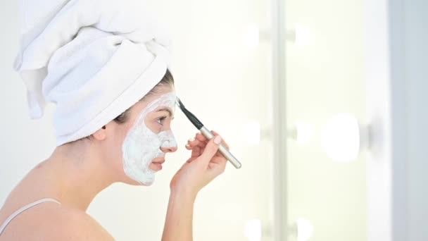 Una mujer con una toalla blanca en la cabeza se aplica una máscara de arcilla azul en la cara con un cepillo. El cuidado de la piel en casa — Vídeos de Stock