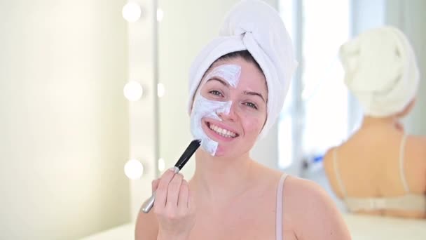 Mooie blanke vrouw met een handdoek op haar haar brengt een klei gezichtsmasker. Zorgen voor schoonheid thuis — Stockvideo