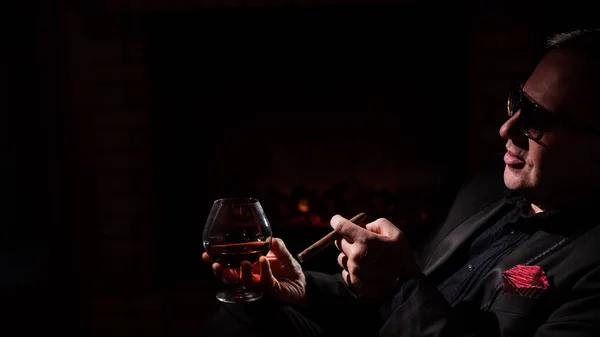 A mature man with glasses drinks brandy and smokes a cigar while sitting in an armchair by the fireplace in the dark. The concept of an elite gentlemens club