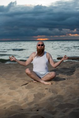 Kızıl sakallı kel bir adam gün batımında plajda yoga yapıyor. Tişörtlü ve güneş gözlüklü komik bir adam deniz kıyısında meditasyon yapıyor..