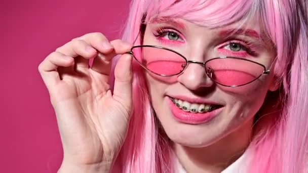 Potret close-up seorang wanita muda dengan kawat gigi di wig merah muda dan kacamata pada latar belakang merah muda. — Stok Video