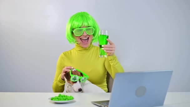 A young woman in a green wig and cheerful glasses drinks beer and bites glazed nuts. A girl sits with a dog at a table and celebrates st patricks day online chatting with friends on a laptop. — Stock Video