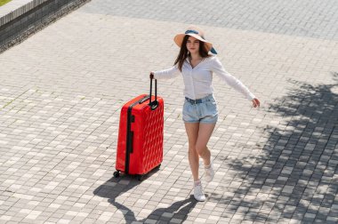 Şapkalı ve şortlu mutlu beyaz kadın açık bir yaşta elinde büyük kırmızı bir bavul tutuyor.