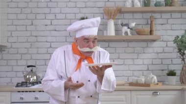 Dost canlısı yaşlı bir şef mutfakta bir tabak tutuyor. Gri saçlı, kalın gri bıyıklı bir adam mutfakta çalışıyor.