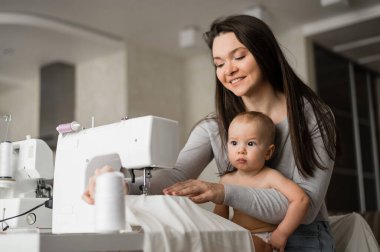 Genç bir kadın evde dikiş dikiyor ve kucağında küçük bir çocuk tutuyor. Annesi küçük oğluna dikiş makinesi dikmeyi öğretiyor.