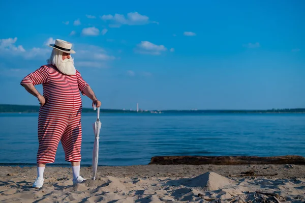 Ein älterer grauhaariger Mann in klassischem Badeanzug und Hut steht mit Regenschirm am Strand und winkt zur Begrüßung mit der Hand. Rentner mit Bart im Urlaub. — Stockfoto