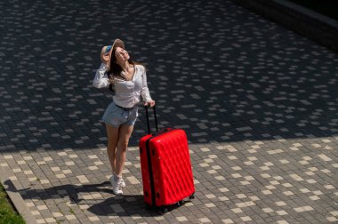 Şapkalı, şortlu, açık havada büyük kırmızı bir bavul tutan mutlu beyaz kadın..