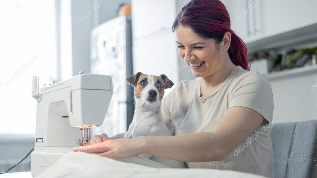 Caucasian woman sews while sitting in the kitchen. Dog Jack Russell Terrier sits on the lap of the owner. Home hobby.