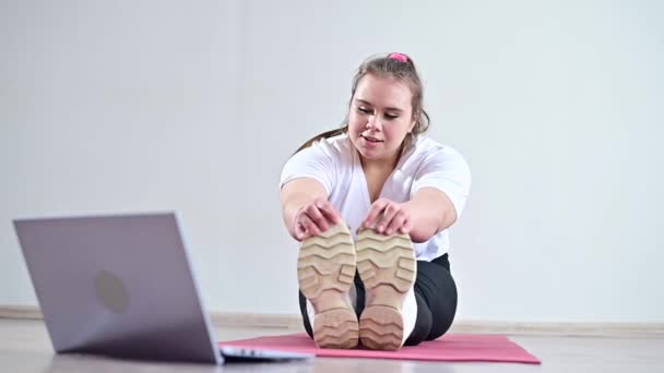 Young caucasian fat woman doing bends on a sports mat and watching a training video on a laptop. A chubby girl doing stretching remotely using video communication — Stock Video