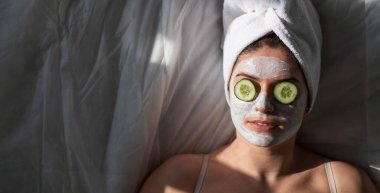 Saçında havlu, yüzünde kilden bir maske ve gözlerinin önünde salatalıklar olan bir kadın çarşafın üzerinde yatıyor.