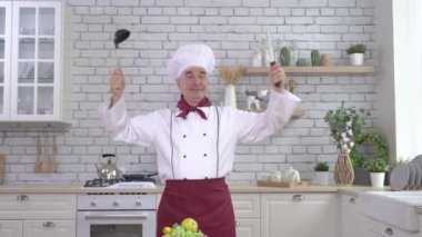 Dost canlısı, yaşlı bir şef aletlerini mutfakta kondüktör gibi sallıyor. Adam bir kepçe tutuyor ve çırpıyor.