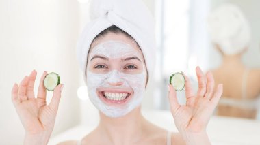 Saçında havluyla ve yüzünde kilden bir maskeyle elinde salatalıkla gezen neşeli bir kadın. Evdeki güzellikle ilgilenmek.