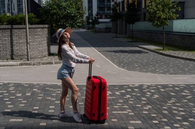 Kafkasyalı güzel esmer kadın, şapkalı, elinde bavuluyla şehir caddesinde poz veriyor..