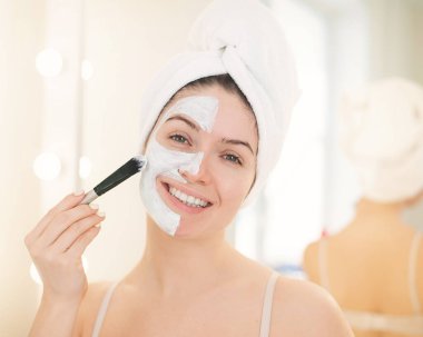 Saçında havlu olan güzel beyaz kadın kilden bir maske takıyor. Evdeki güzellikle ilgilenmek.