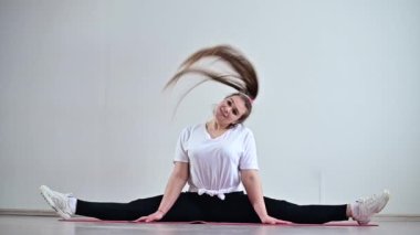 Genç, esnek, şişman bir kadın enine bir sicimin içinde oturur ve saçını beyaz bir arka plana doğru sallar. Yavaş çekim