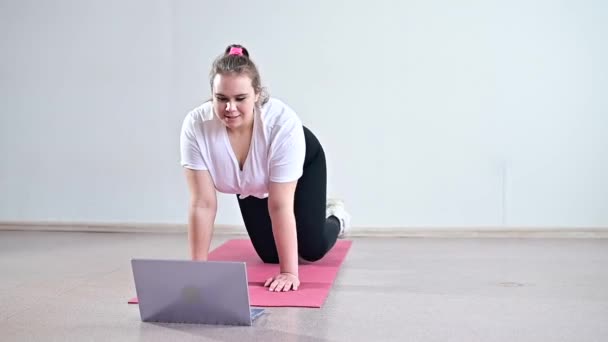 Ung kvinna som motionerar hemma. Flickan är engagerad i fitness hemma på mattan och tittar på en träningsvideo på en bärbar dator — Stockvideo