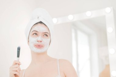 Saçında havlu olan güzel beyaz kadın kilden bir maske takıyor. Evdeki güzellikle ilgilenmek.