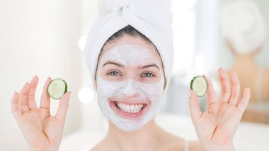 Saçında havluyla ve yüzünde kilden bir maskeyle elinde salatalıkla gezen neşeli bir kadın. Evdeki güzellikle ilgilenmek.