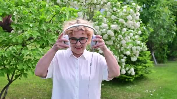 快乐的老妇人走在盛开的苹果树的公园里，戴上耳机。一位女性退休人员正在花园里度过一个温暖的夏天，听着音乐。老一代人. — 图库视频影像