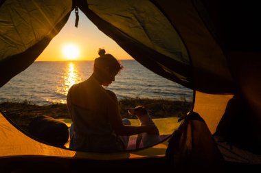 Gün batımında bir kadın ve bir köpek doğadaki bir çadırda dinleniyorlar. Kız ve Jack Russell Terrier nehir kıyısında kamp kurmuşlar.