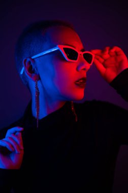 Stüdyoda kısa erkek saç kesimli, güneş gözlüklü, neon ışıklı genç bir kadının portresi.