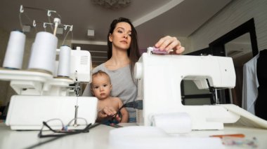 Genç bir kadın evde dikiş dikiyor ve kucağında küçük bir çocuk tutuyor. Annesi küçük oğluna dikiş makinesi dikmeyi öğretiyor.