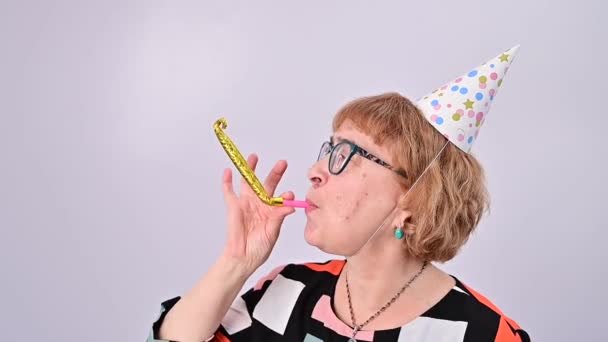 Feliz anciana con gorra de cumpleaños haciendo sonar un silbato sobre un fondo blanco — Vídeos de Stock