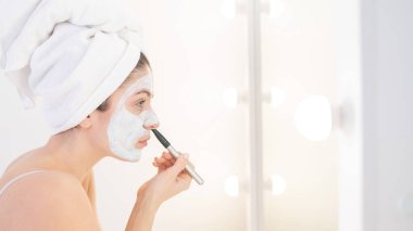 Saçında havlu olan güzel beyaz kadın kilden bir maske takıyor. Evdeki güzellikle ilgilenmek.