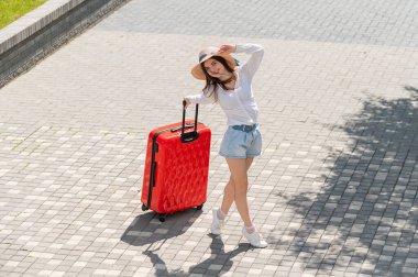 Şapkalı ve şortlu mutlu beyaz kadın açık bir yaşta elinde büyük kırmızı bir bavul tutuyor.
