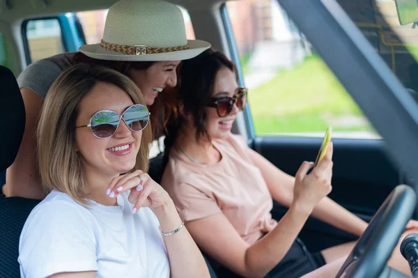 Três namoradas felizes fazem uma viagem. Mulheres Andar de carro, olhar para o telefone e rir. — Fotografia de Stock