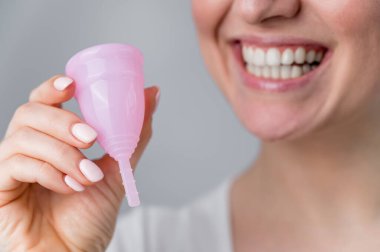 Beyaz arka planda pembe adet bardağı tutan gülümseyen beyaz kadının yakın plan portresi. Kritik günlerde tampon ve pedlere alternatif.