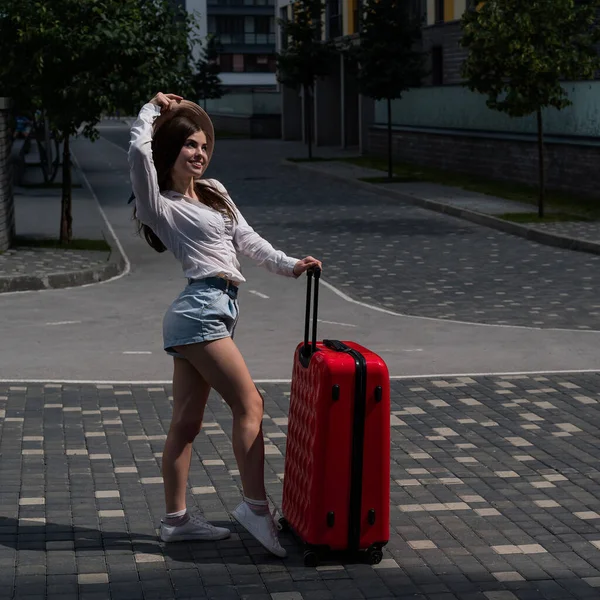 Schöne kaukasische brünette Frau mit Hut posiert mit einem Koffer auf einer Straße der Stadt. — Stockfoto