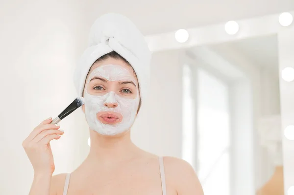 Hermosa mujer caucásica con una toalla en el pelo se aplica una mascarilla de barro. Cuidar de la belleza en casa — Foto de Stock